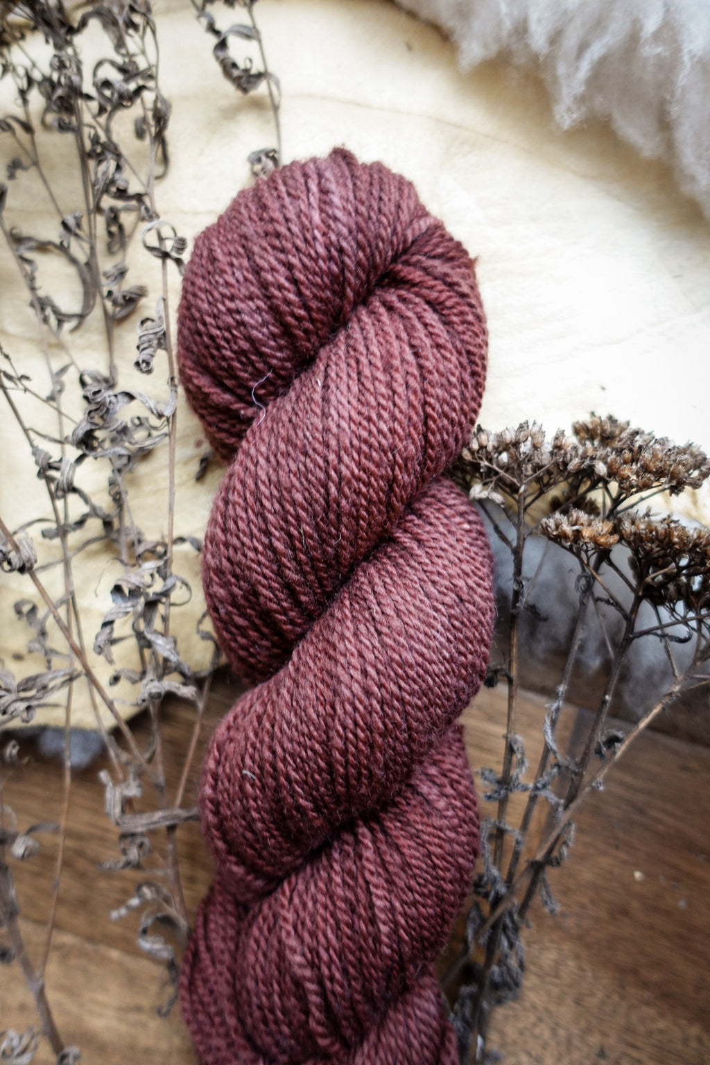 A DK weight skein of natural fiber yarn has been hand dyed a reddish pink. It lays on a tabletop next to dried flowers.