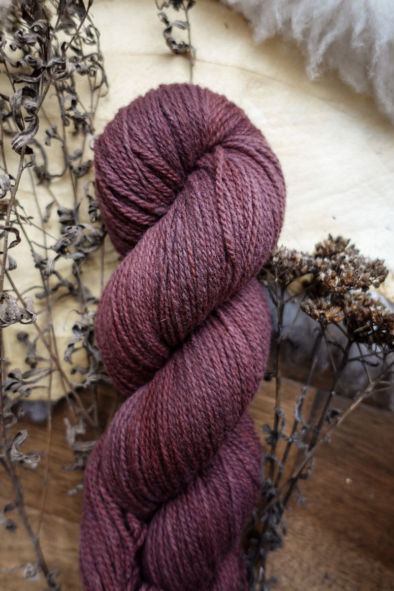 A natural fiber skein of reddish pink yarn lays on a tabletop, next to dried flowers.