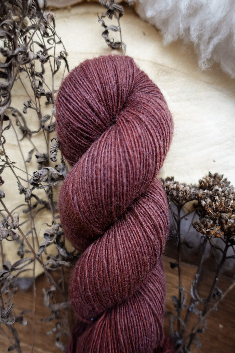 A reddish pink skein of hand dyed, fingering weight yarn lays on a tabletop next to dried flowers.
