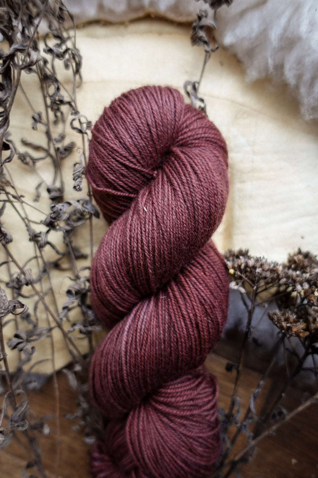 A fingering weight skein of natural fiber yarn has been hand dyed a reddish pink. It lays on a tabletop next to dried flowers.