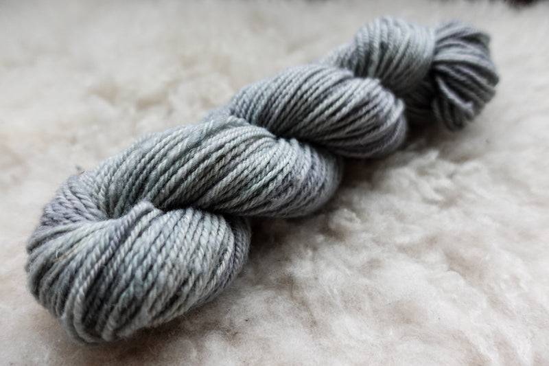 A worsted weight, natural fiber skein of yarn has been hand dyed a light grey. It lays on a sheepskin rug.