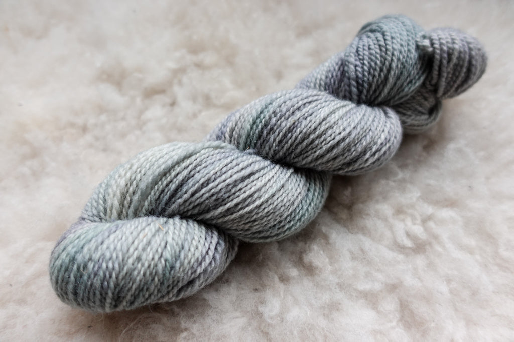 A natural fiber skein of yarn has been hand dyed light grey. It lays on a sheepskin rug.