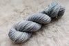 A sport weight skein of natural fiber yarn has been hand dyed a light grey. It lays on a wool background.