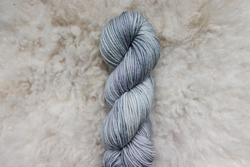 A natural fiber skein of hand dyed, light grey yarn lays on a sheepskin rug, pictured from above.