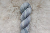 A light grey skein of hand dyed yarn lays on a sheepskin rug, pictured from above.