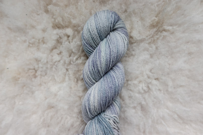 A light grey skein of hand dyed yarn lays on a sheepskin rug, pictured from above.