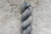 A naturally dyed skein of light grey yarn lays on a sheepskin rug, pictured from above.