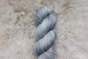 A hand dyed skein of light grey yarn lays on a sheepskin rug, pictured from above.