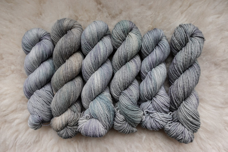 Six skeins of yarn, all different bases, have been hand dyed a light grey. They lay on a sheepskin rug.
