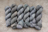 Six skeins of yarn, all different bases, have been hand dyed a light grey. They lay on a sheepskin rug.