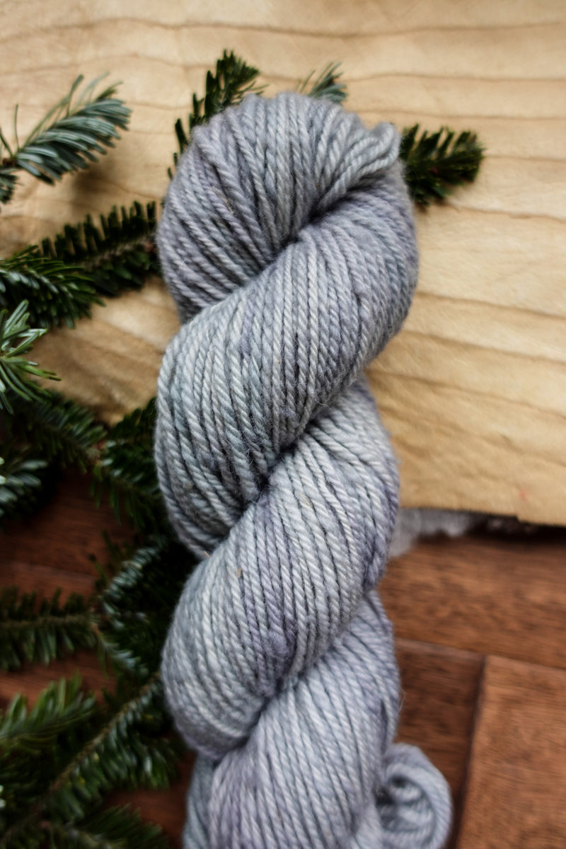 A light grey skein of hand dyed yarn lays on a tabletop next to fir branches.