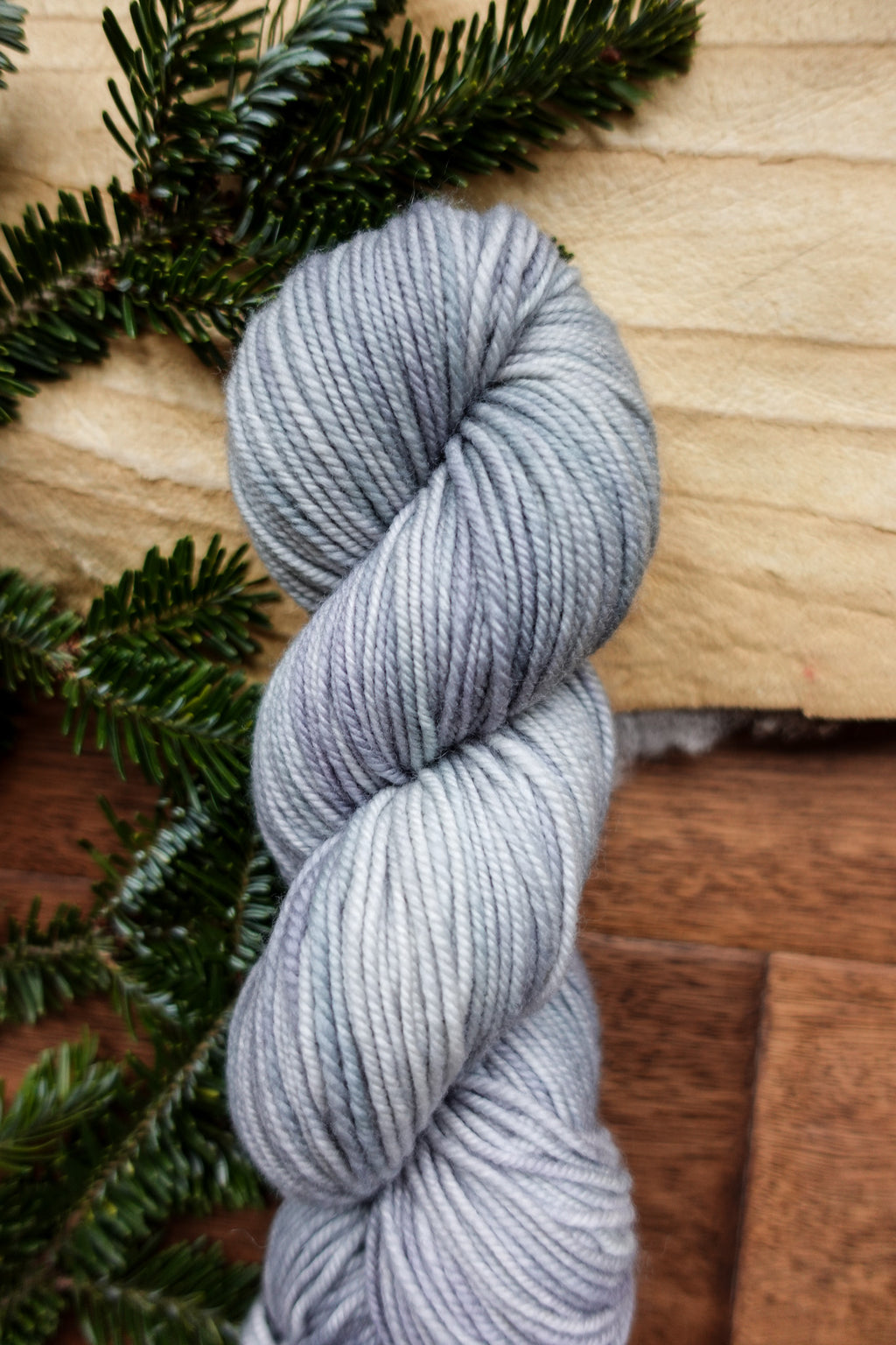 A worsted weight skein of yarn has been hand dyed a light grey. It lays on a tabletop next to fir branches.