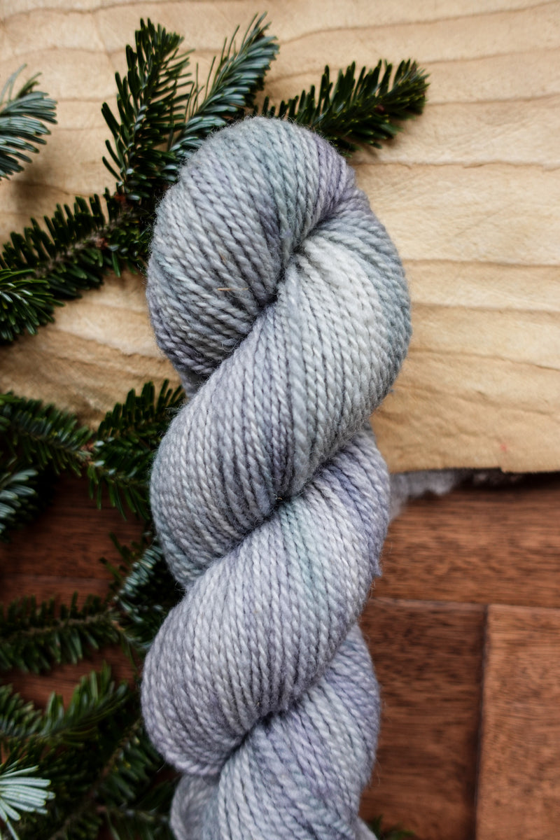 A light grey skein of hand dyed, DK weight yarn lays on a tabletop. Fir branches can be seen in the background.