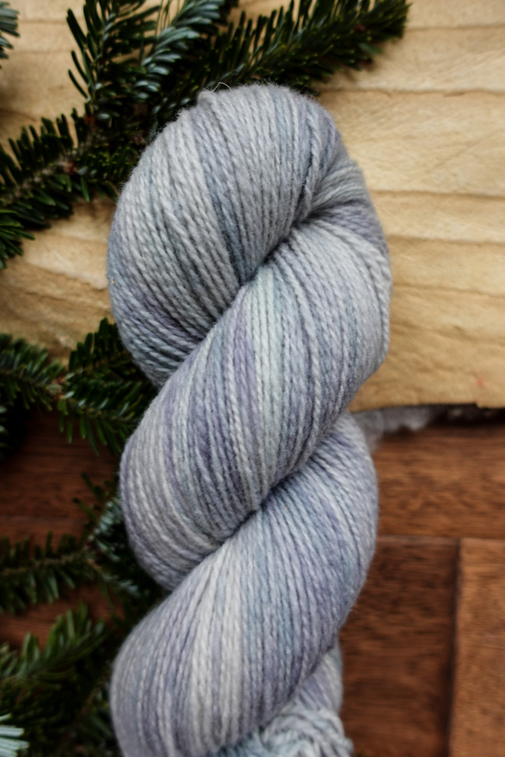 A hand dyed skein of light grey yarn lays on a tabletop, next to fir branches.