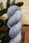 A skein of single ply, fingering weight yarn has been hand dyed a light grey. It lays on a tabletop next to fir branches.
