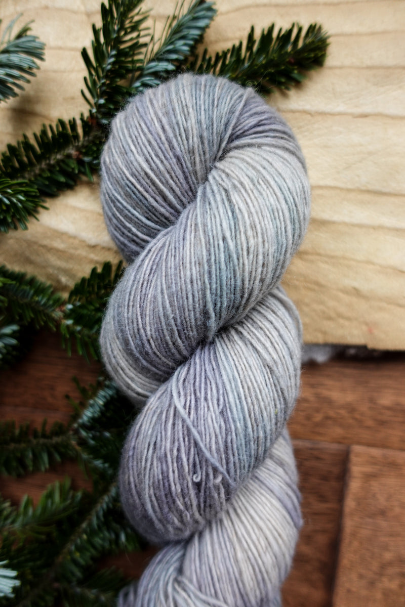 A skein of single ply, fingering weight yarn has been hand dyed a light grey. It lays on a tabletop next to fir branches.