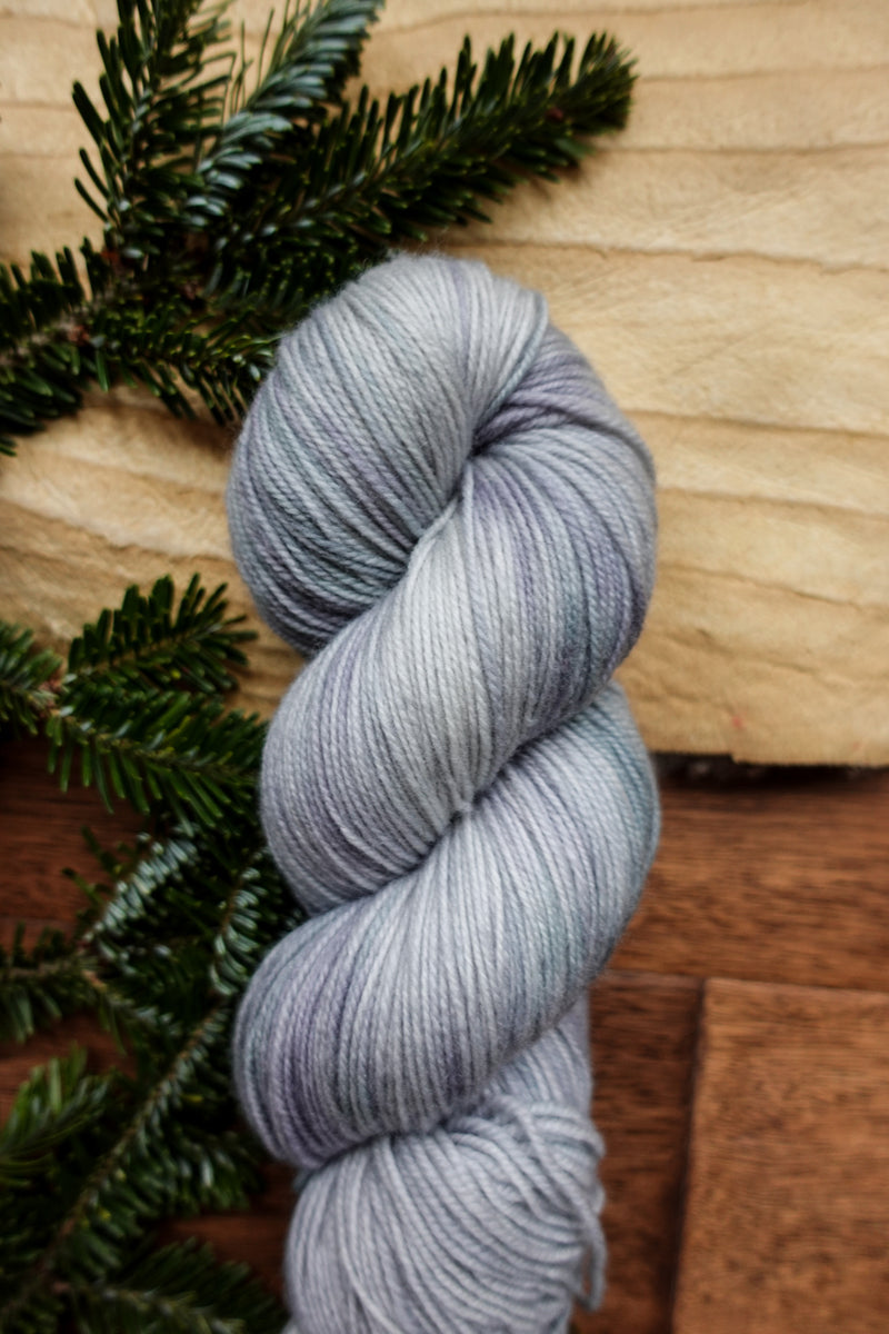 A sock weight skein of yarn has been hand dyed a light grey. It lays on a tabletop next to fir branches.