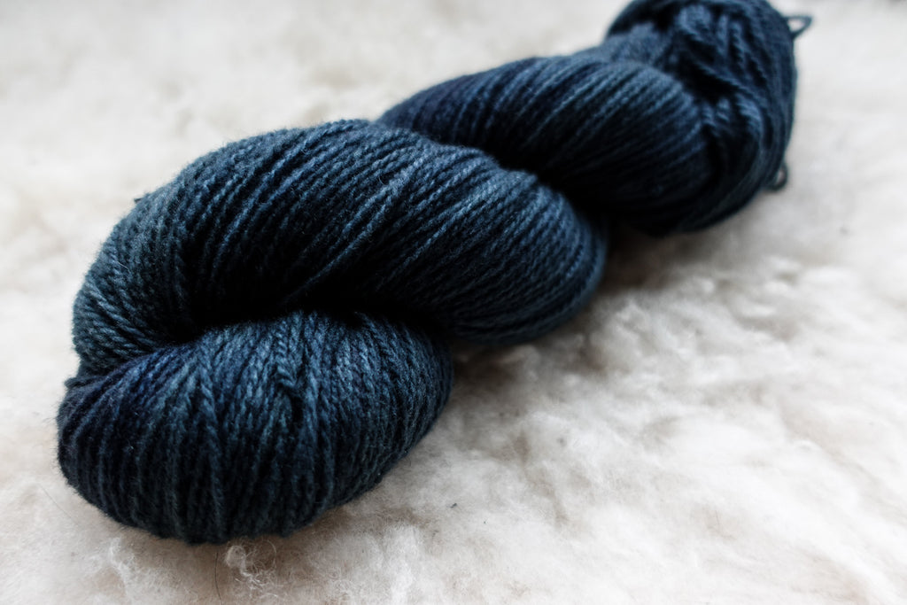 A sport weight skein of natural fiber yarn has been hand dyed a deep blue. It lays on a wool background.