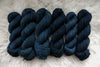 Six skeins of yarn, all different bases, have been hand dyed a deep blue. They lay on a sheepskin rug.