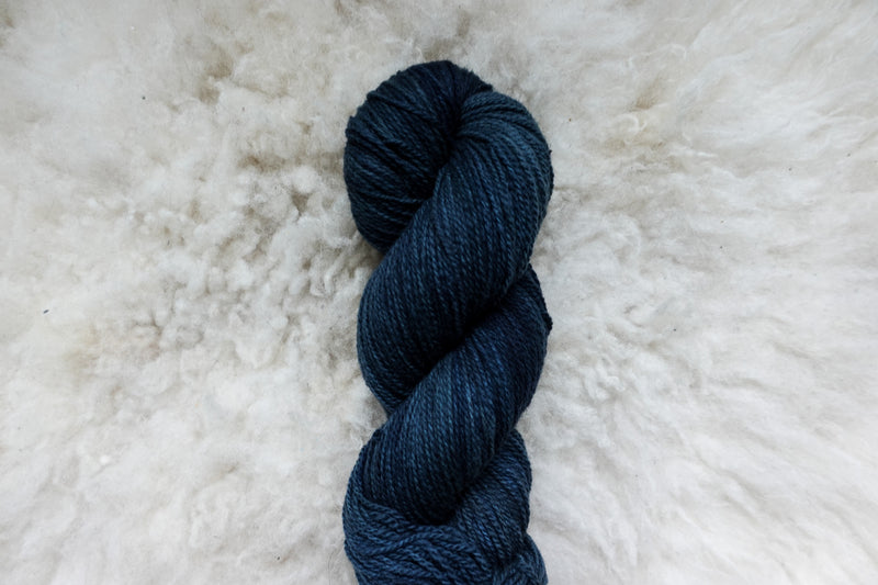 Pictured from above, a natural fiber skein of deep blue yarn lays on a sheepskin rug.