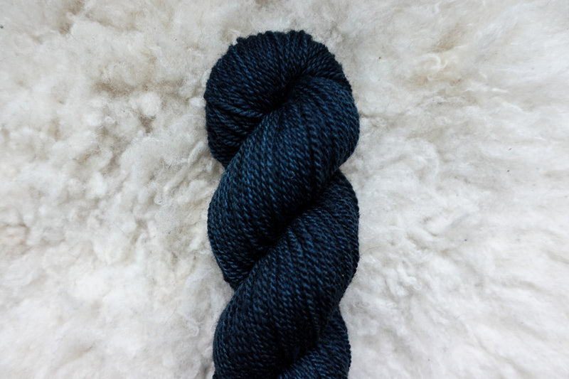 A natural fiber skein of yarn has been hand dyed a deep blue. It lays on a sheepskin rug, pictured from above.