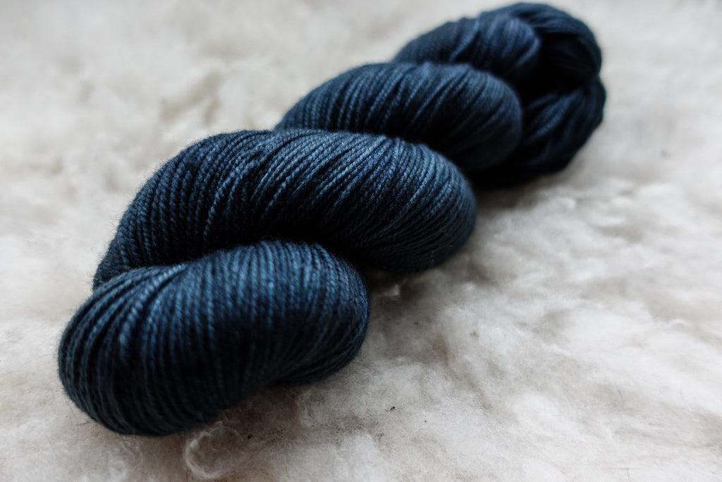 A natural fiber skein of yarn has been hand dyed a deep blue. It lays on a sheepskin rug.
