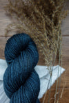 A skein of worsted weight yarn has been hand dyed deep blue. It lays on a tabletop next to dried grasses.