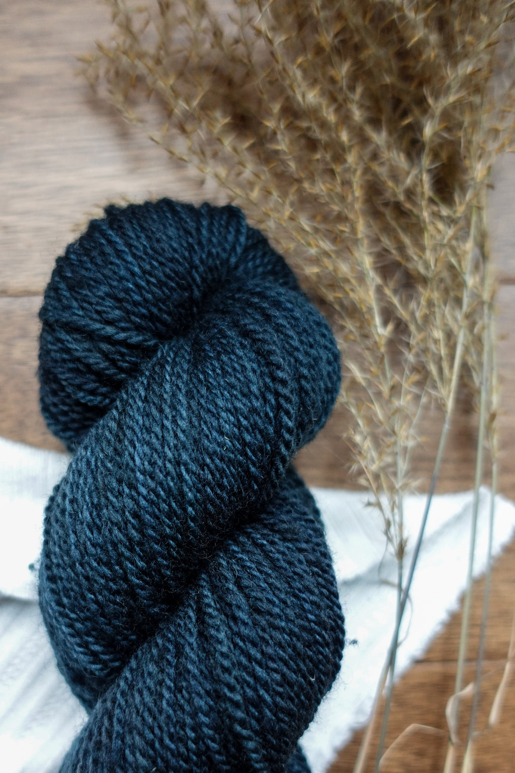 A DK weight skein of deep blue yarn lays on a tabletop next to dried grasses.