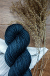 A skein of sock weight yarn has been hand dyed a deep blue. It lays on a tabletop next to dried grasses.