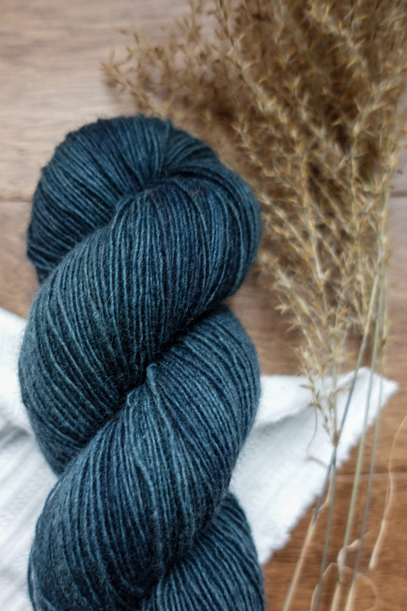 A skein of single ply yarn has been naturally dyed deep blue. It lays on a tabletop next to dried grasses.