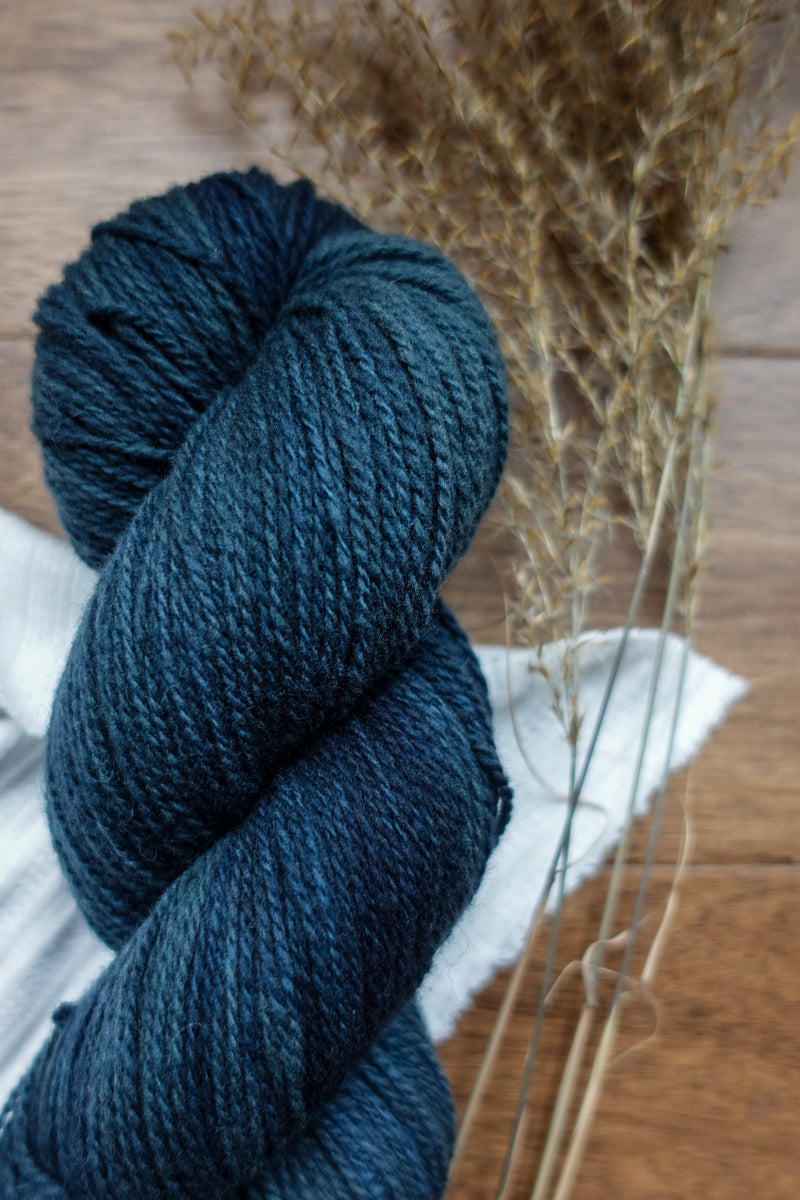 A sport weight skein of yarn has been dyed a deep blue. It lays on a tabletop next to dried grasses.