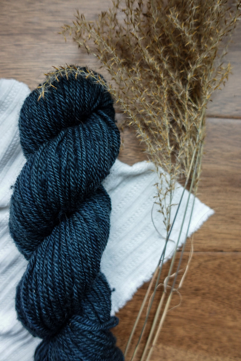 A deep blue skein of worsted weight yarn lays on a tabletop next to dried grasses.