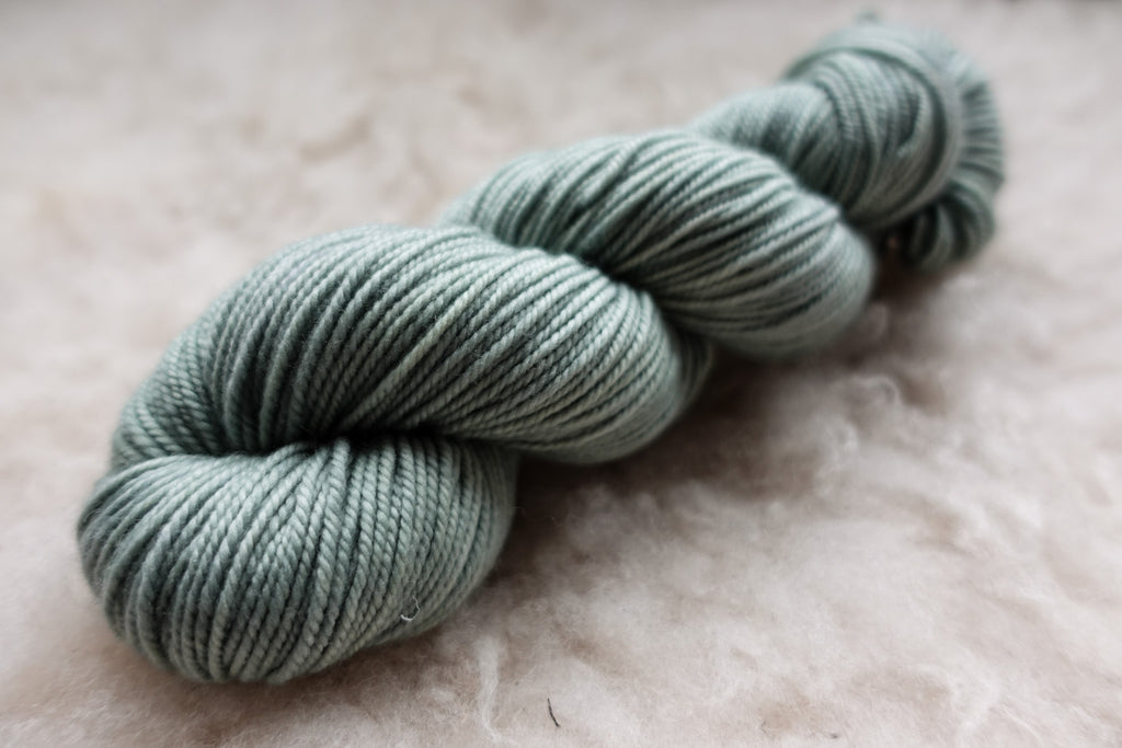 A worsted weight skein of natural fiber yarn has been hand dyed a light grey. It lays on a wool background.