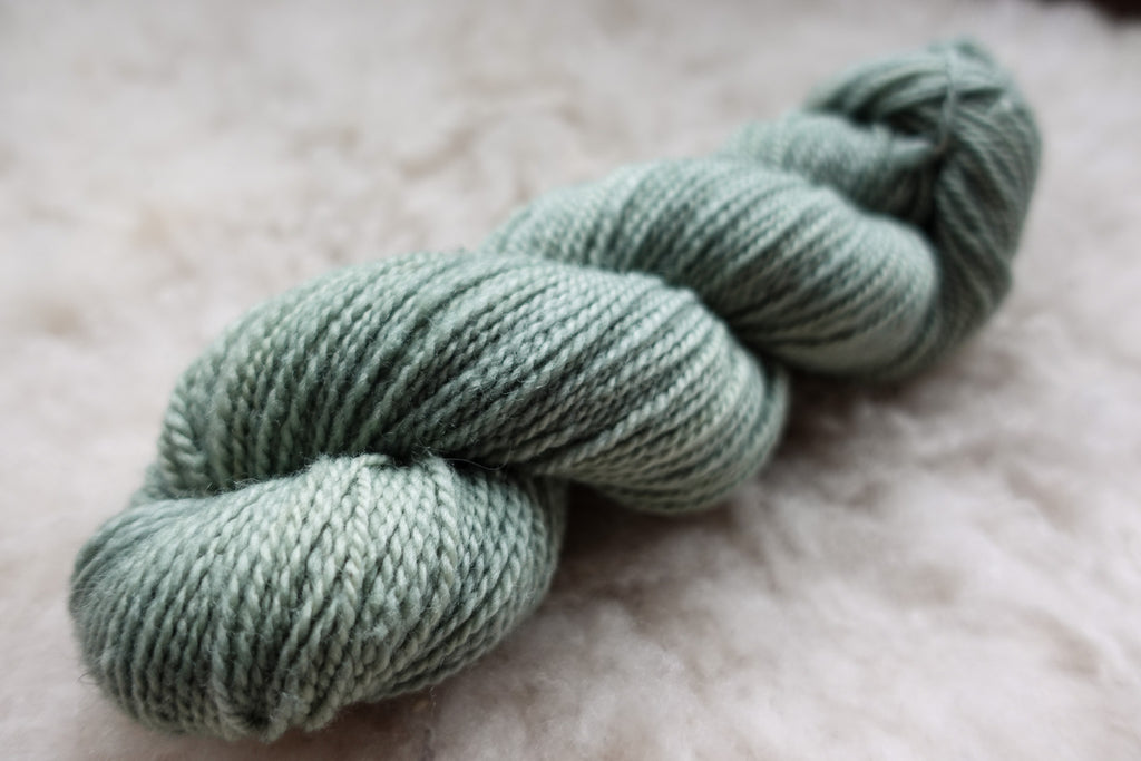 A DK weight skein of natural fiber yarn has been hand dyed a light grey. It lays on a wool background.