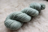 A sport weight skein of natural fiber yarn has been hand dyed a light grey. It lays on a wool background.