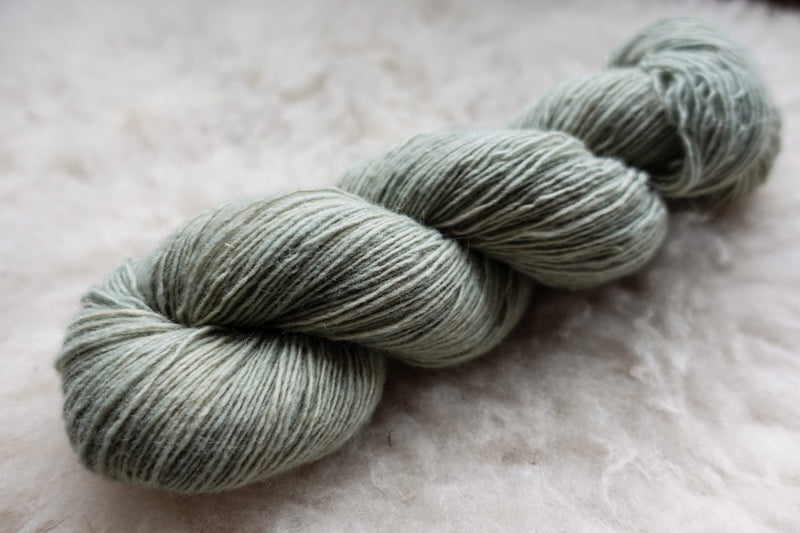 A fingering weight skein of natural fiber yarn has been hand dyed a light grey. It lays on a wool background.