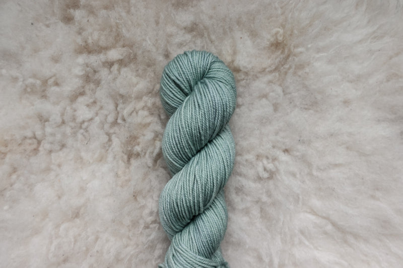 A skein of yarn has been naturally dyed a light grey. It lays on a sheepskin rug and is pictured from above.