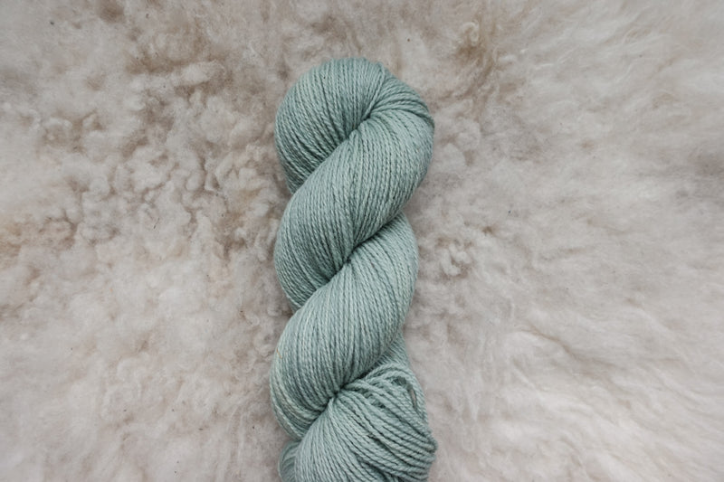 A light grey skein of naturally dyed yarn lays on a sheepskin, and is pictured from above.