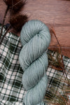 A hand dyed skein of worsted weight yarn lays on a tabletop next to dried grasses and a plaid cloth.