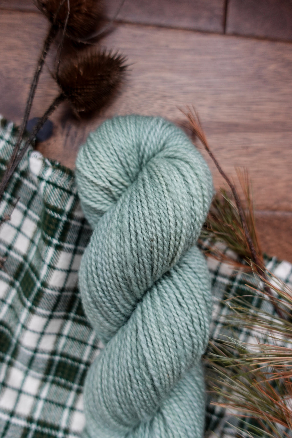 A skein of naturally dyed, light grey yarn lays on a plaid cloth. Dried branches can be seen in the background.
