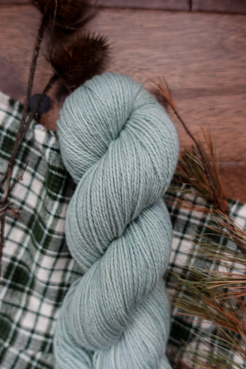 A skein of sport weight yarn has been hand dyed a light grey. It lays on a plaid cloth, with dried branches in the background.