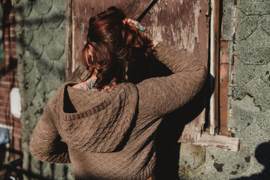 Elizabeth, seen from behind, holds up her hair to show off the hood on her hand knit, gansey style sweater. She stands in front of an old brick wall.