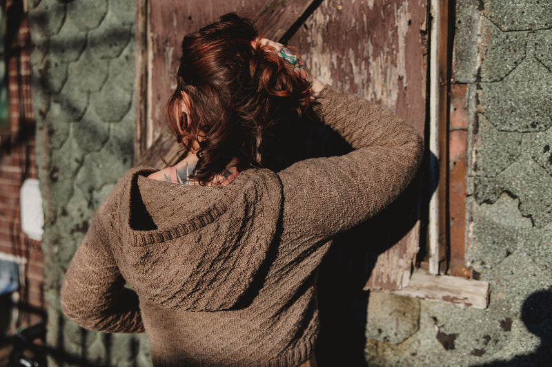 Elizabeth, seen from behind, holds up her hair to show off the hood on her hand knit, gansey style sweater. She stands in front of an old brick wall.