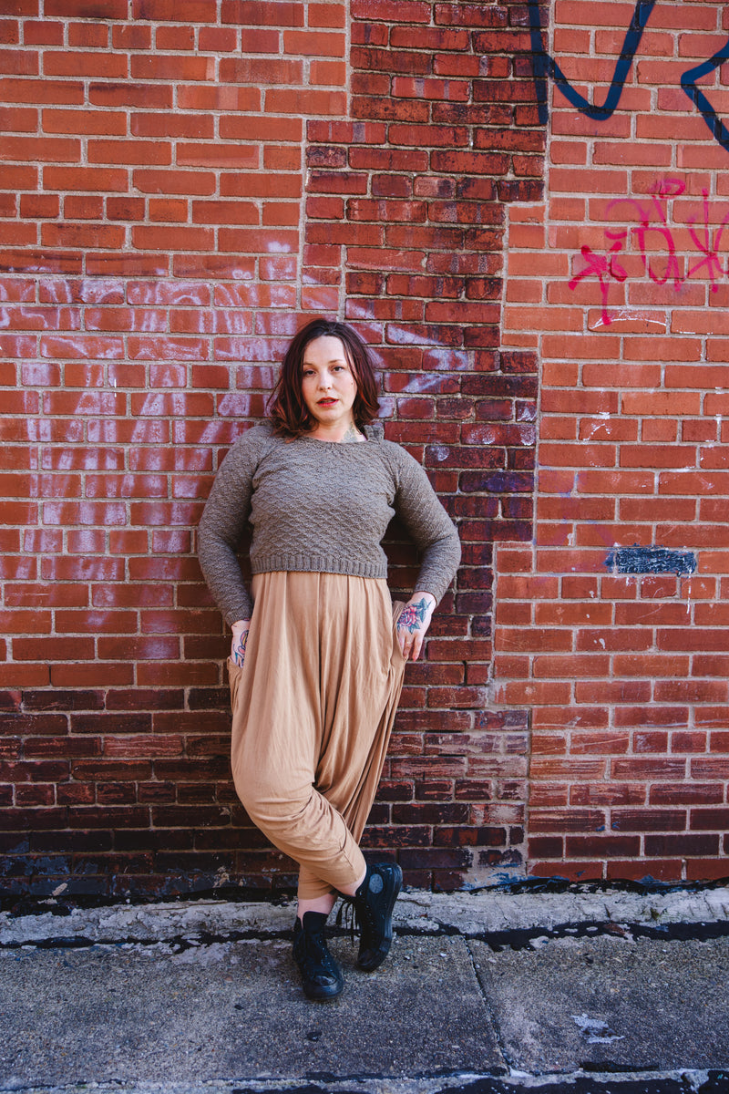 Elizabeth, standing in front a brick wall, wears a light brown cropped sweater, hand knit with a diamond stitch pattern. She's paired it with black high tops and tan pants.