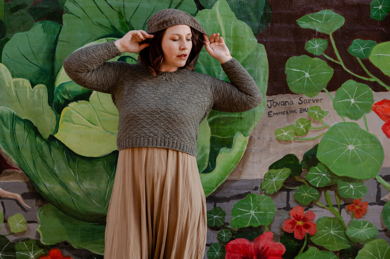 Elizabeth pulls up the hood of her hand knit, light brown sweater. She looks to the side, and stands in front of a wall mural of flowers and leaves.