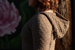 Seen from the side, Elizabeth wears a light brown hoodie, hand knit with a diamond stitch pattern. The sun is at her back, illuminating the ribbed edge of the hood.