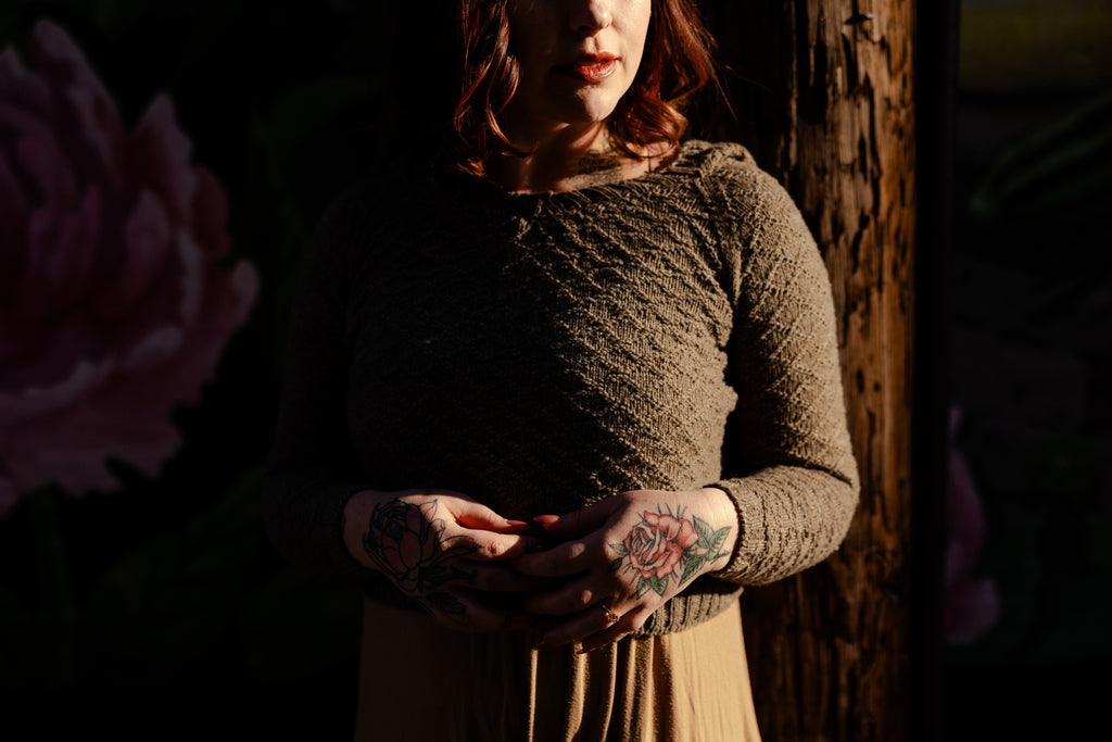Elizabeth, a white woman with brown hair, wears a light brown hooded sweater, hand knit with a gansey diamon stitch. She is partially in shadow, her head turned towards the sunlight.