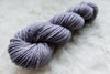 A worsted weight skein of natural fiber yarn has been hand dyed a light purple. It lays on a wool background.