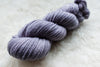 A skein of sport weight, natural fiber yarn has been hand dyed a light purple. It lays on a wool background.