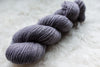 A single ply skein of fingering weight yarn has been hand dyed a light purple. It lays on a wool background.